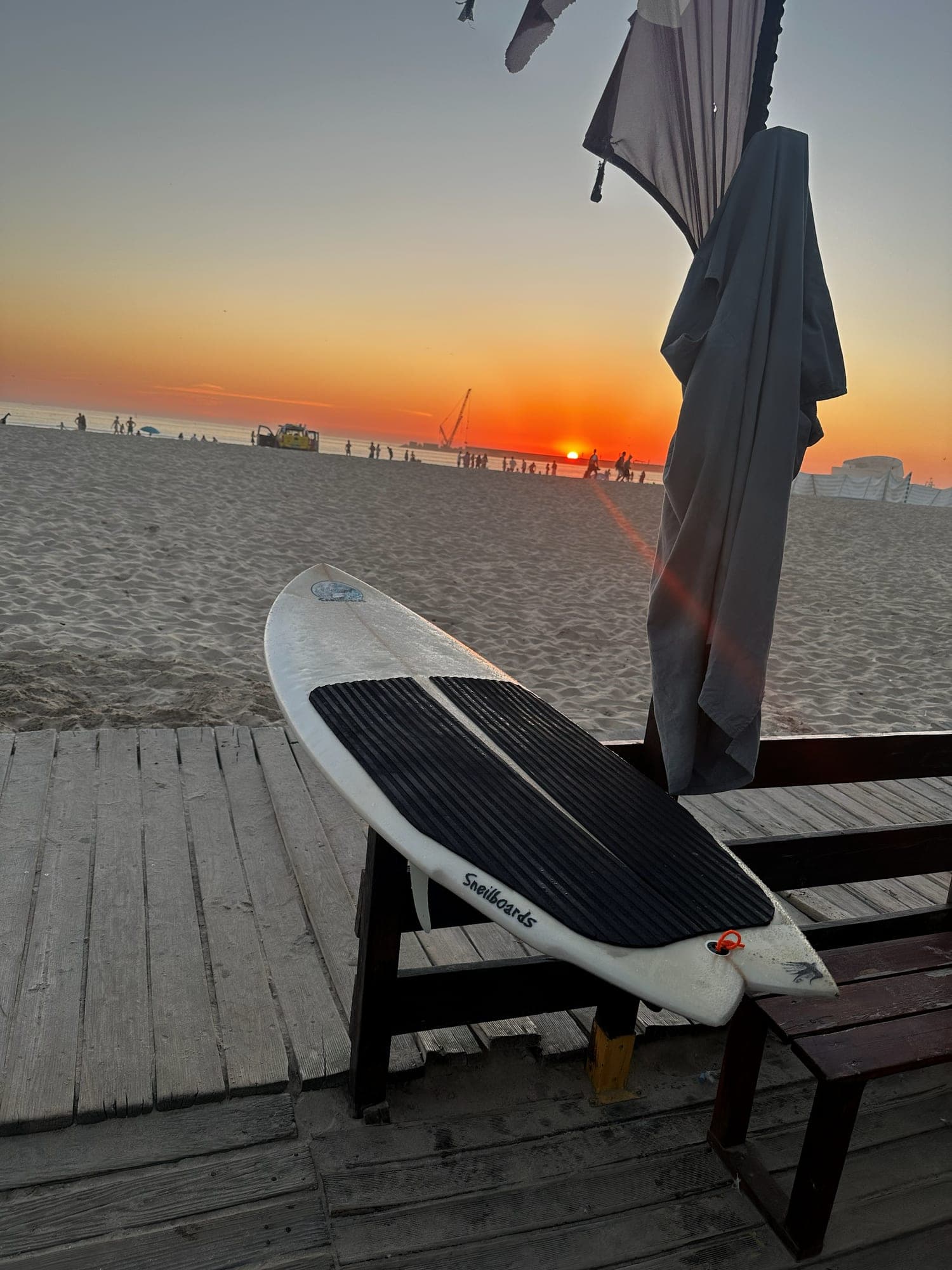 A surfboard leaning on a wooden bench on a beach boardwalk at sunset, with a wetsuit hanging beside it