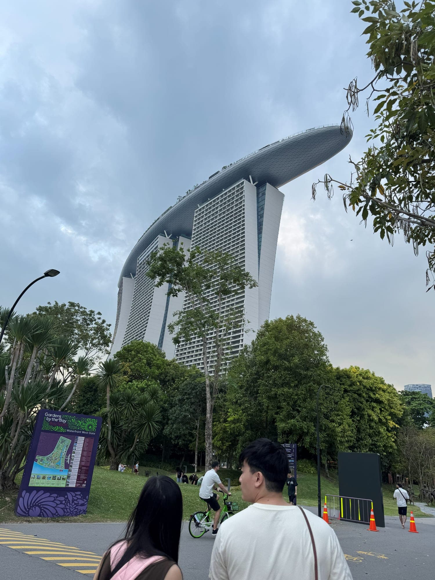 Marina Bay Sands' three towers and rooftop deck seen from a path in Gardens by the Bay under a cloudy sky