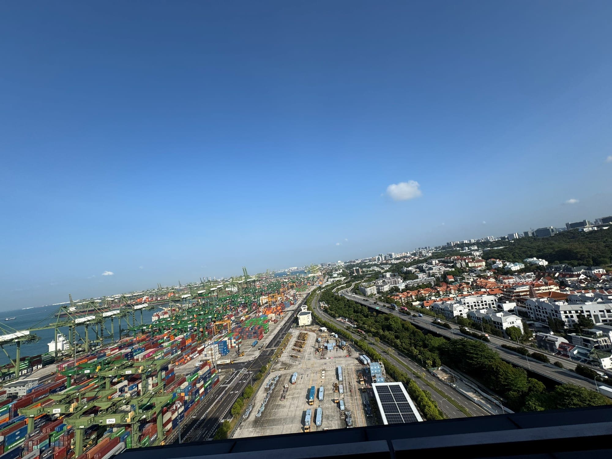 A view from above over a container terminal with rows of cranes and stacked containers, next to a motorway and housing