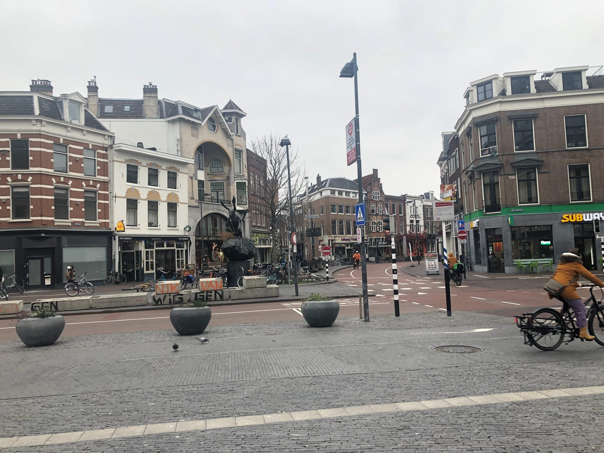 A square in Utrecht's city centre on a grey day, with brick town houses, a dark statue, large plant pots and a cyclist on a red bike lane