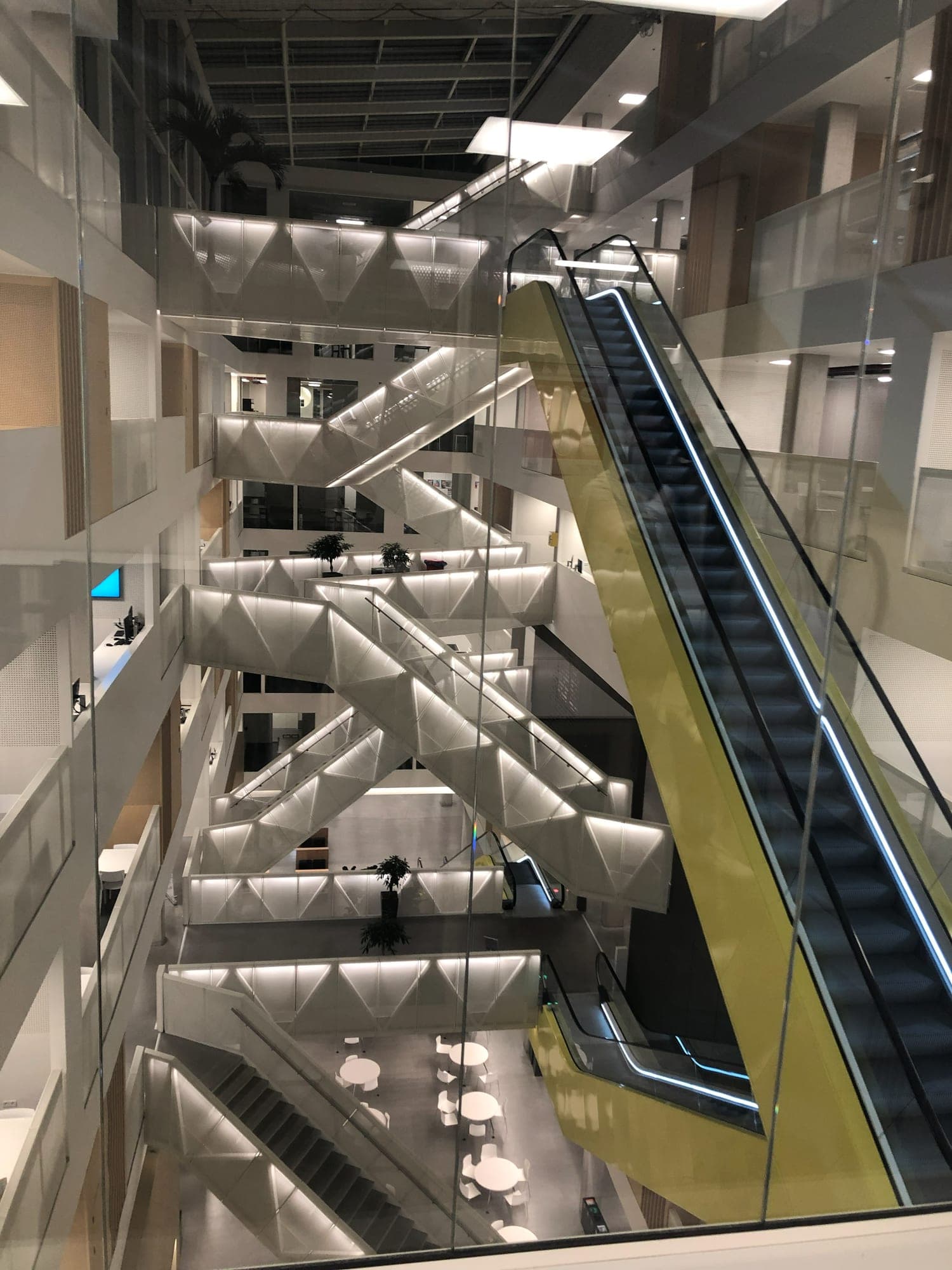 A long escalator in a yellow casing rising through an atrium of white staircases, seen through a glass wall