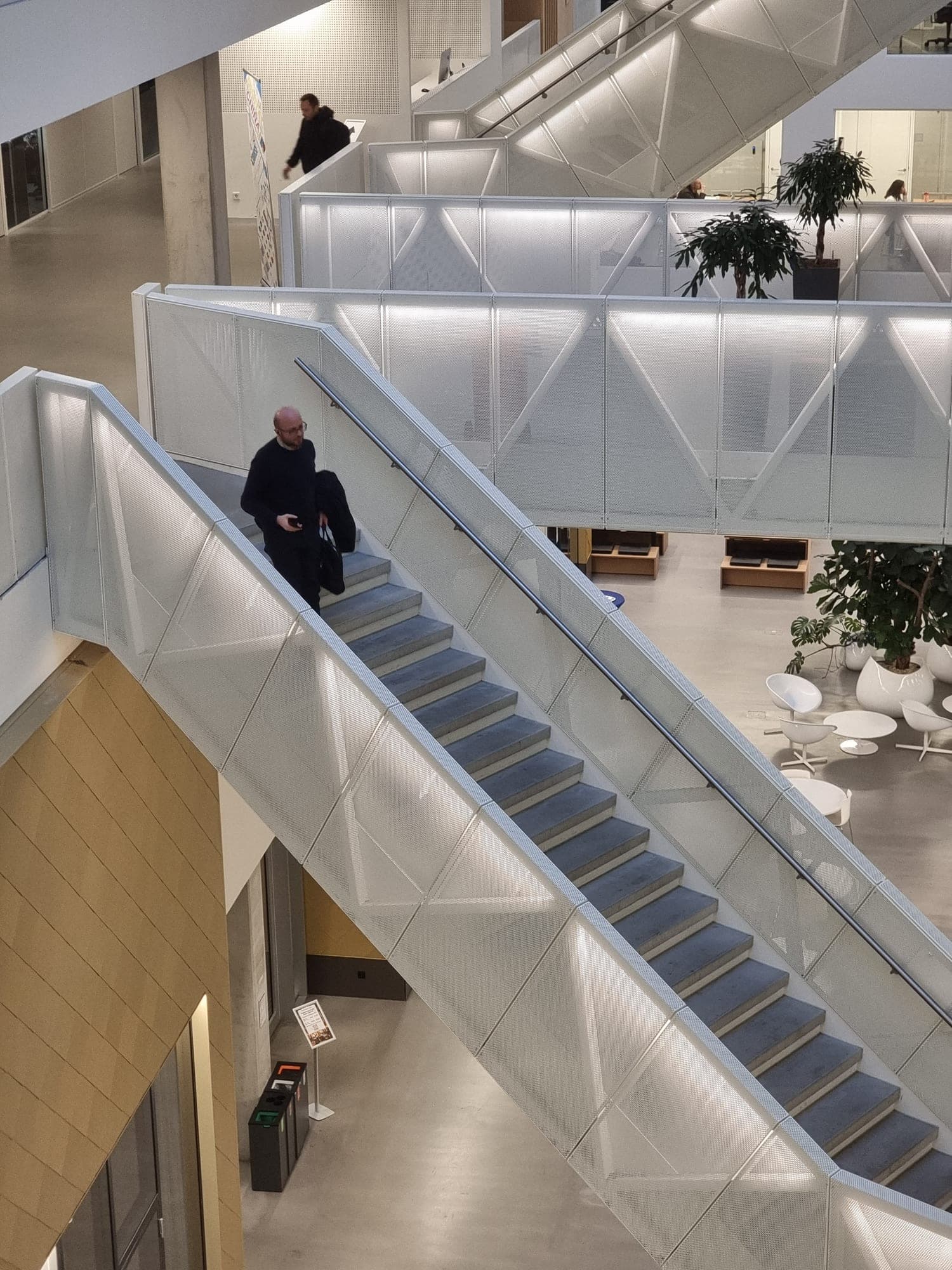 A man walking down a wide staircase with white translucent balustrades in a bright atrium with plants and white chairs below