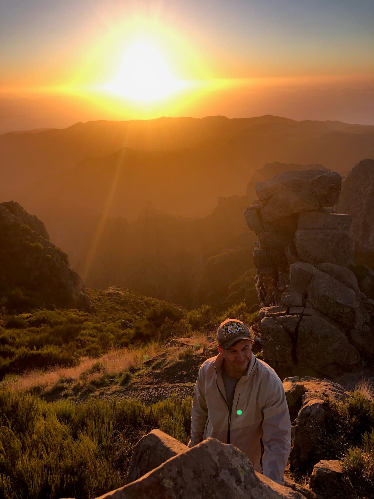 A hiker in a cap climbing between rocks as the sun rises over the mountain ridges of Madeira