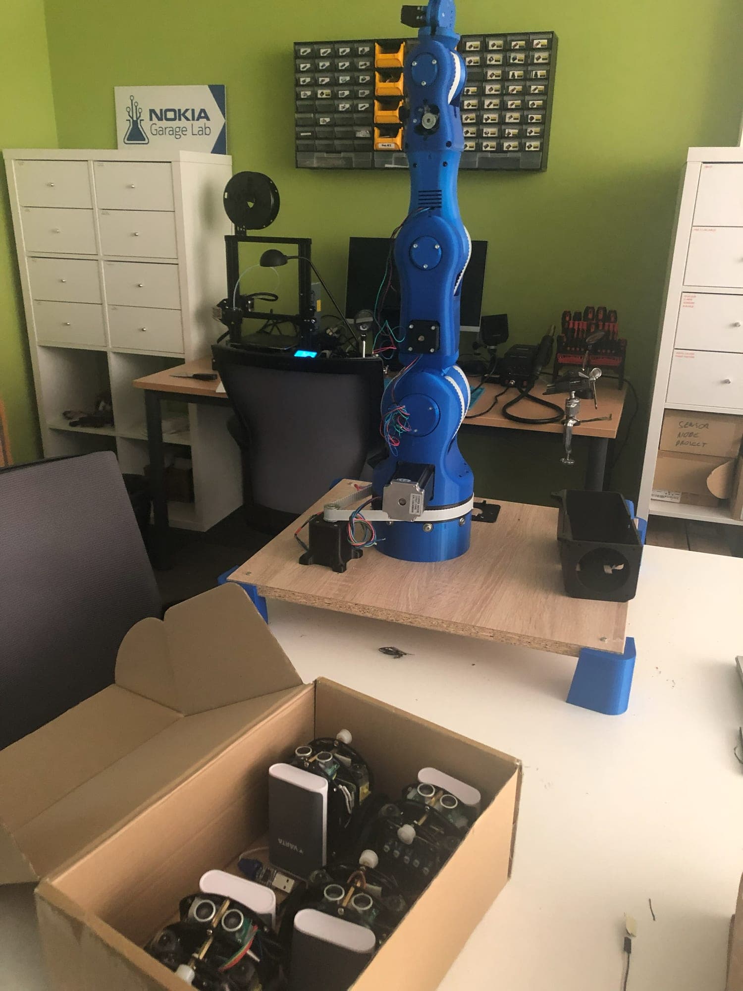 A box of small robot cars on a desk in the Nokia Garage Lab, with a blue 3D-printed robot arm, a 3D printer and component drawers behind