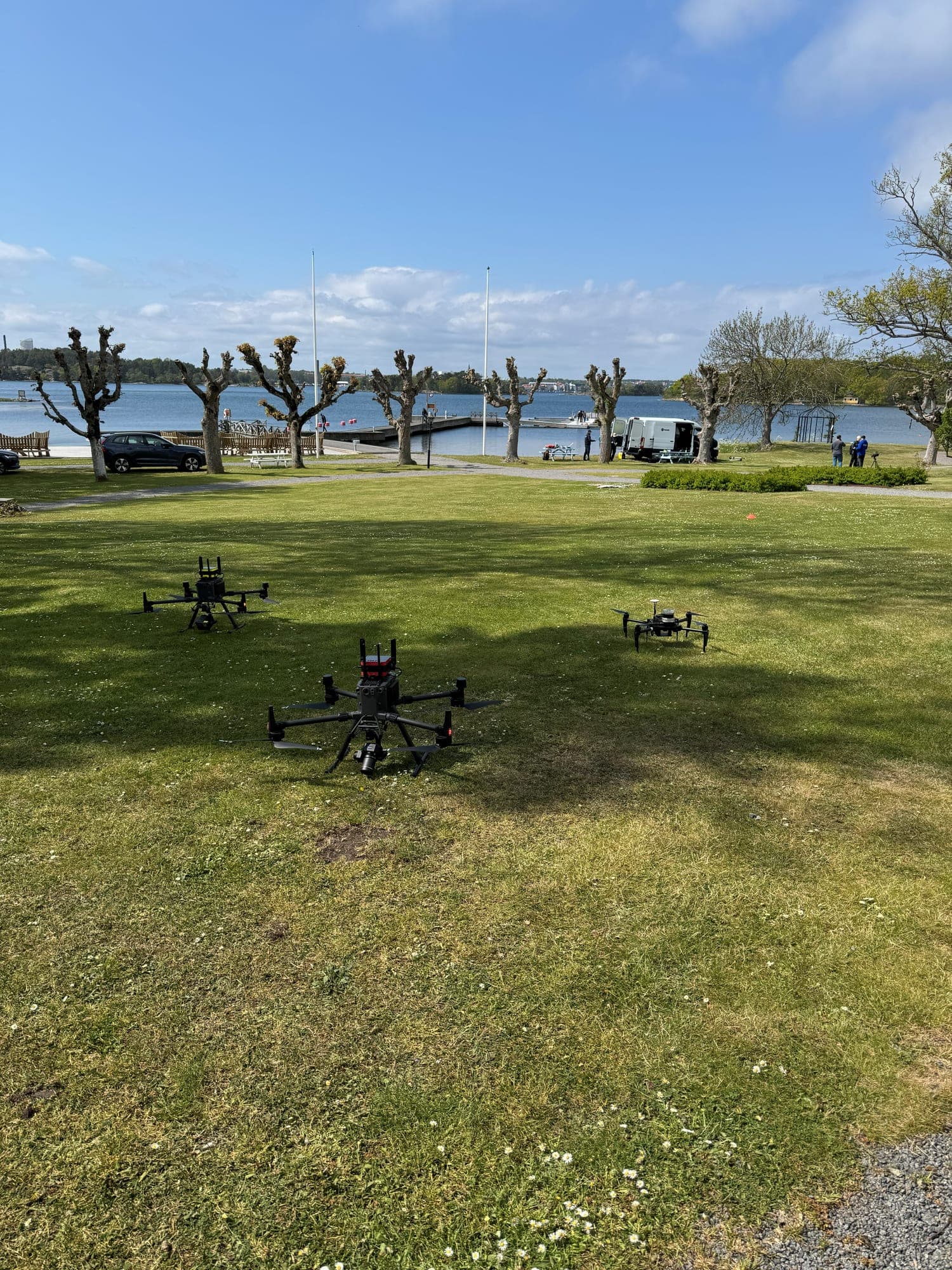 Three quadcopter drones on a lawn by the water, with a jetty and a van in the background